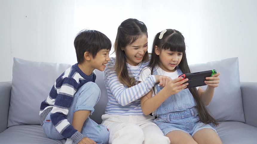 Three children sit on a sofa in a bright, softly lit room, happily playing on a handheld gaming device.