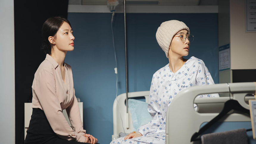 Two women sit in a hospital room; one in a patient gown and hat looks concerned, while the other listens attentively under soft, focused lighting.