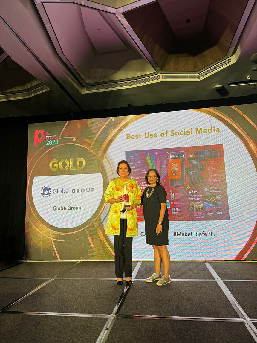 A couple of women standing indoors in front of a sign that reads "Best Use of Social Media GOLD Globe GROUP #MakeITSafePH."