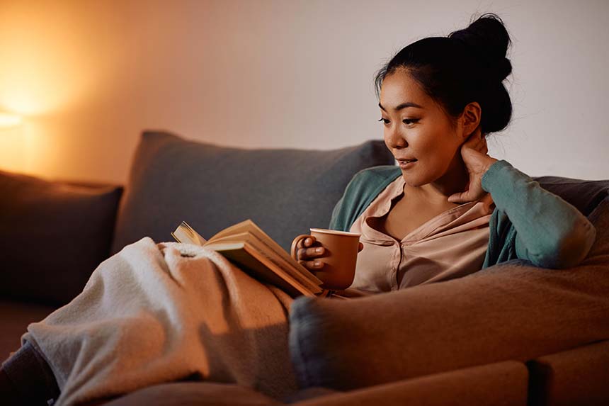A woman relaxes on a sofa under warm, dim lighting, wrapped in a blanket, holding a mug, and reading a book.