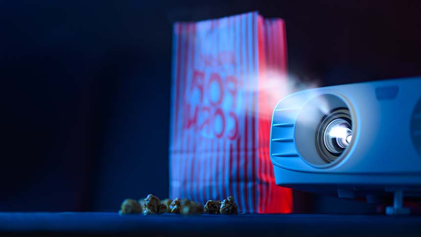A projector beams light toward a screen, with a red-and-white striped popcorn bag and scattered popcorn in a dimly lit setting.