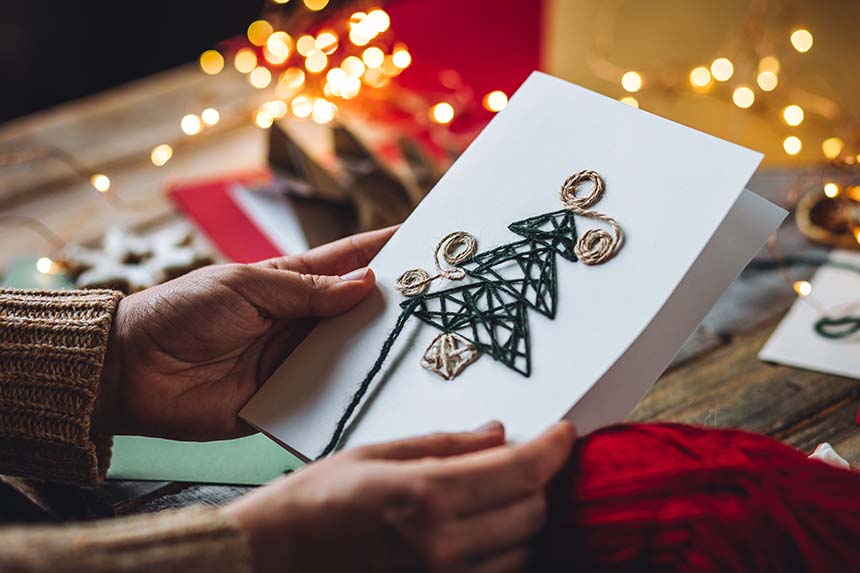 A person holding a handmade Christmas card decorated with string art, shaped like a Christmas tree, surrounded by festive lights and a rustic table setting.