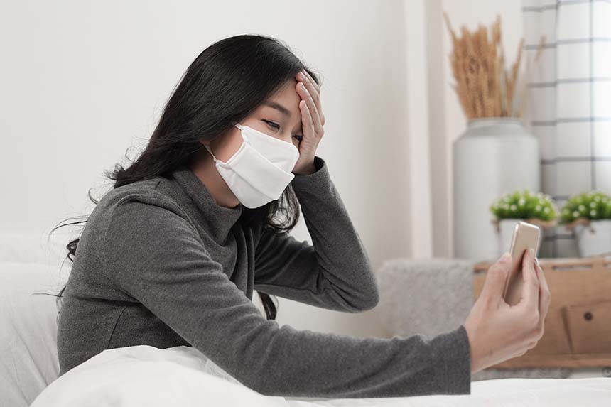 A woman wearing a face mask, sitting in bed, holding her head while looking at her smartphone, suggesting illness or a teleconsultation.