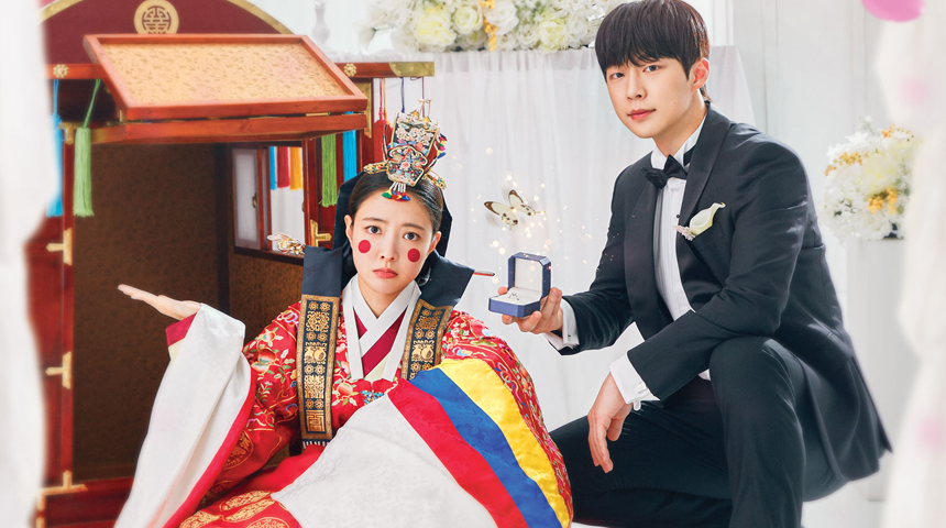 A man in a tuxedo kneels with a ring box, while a woman in traditional Korean attire looks surprised, sitting beside a colorful wedding palanquin in a bright, festive setting.