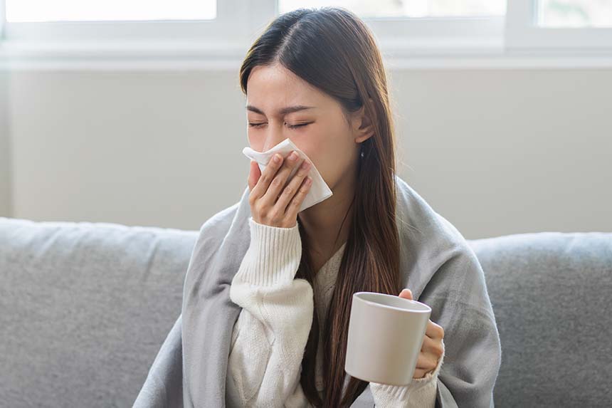 A woman sitting on a couch with a blanket, sneezing into a tissue while holding a mug, conveying a sense of illness or cold weather.